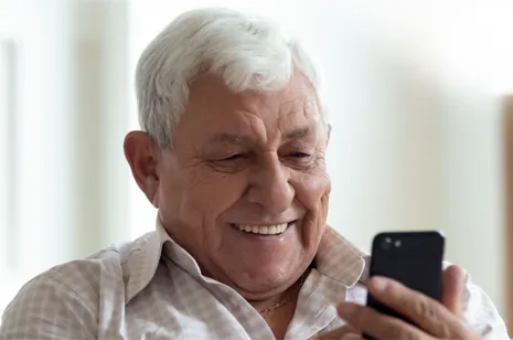 Smiling elderly man in a checkered shirt looking at a black smartphone.