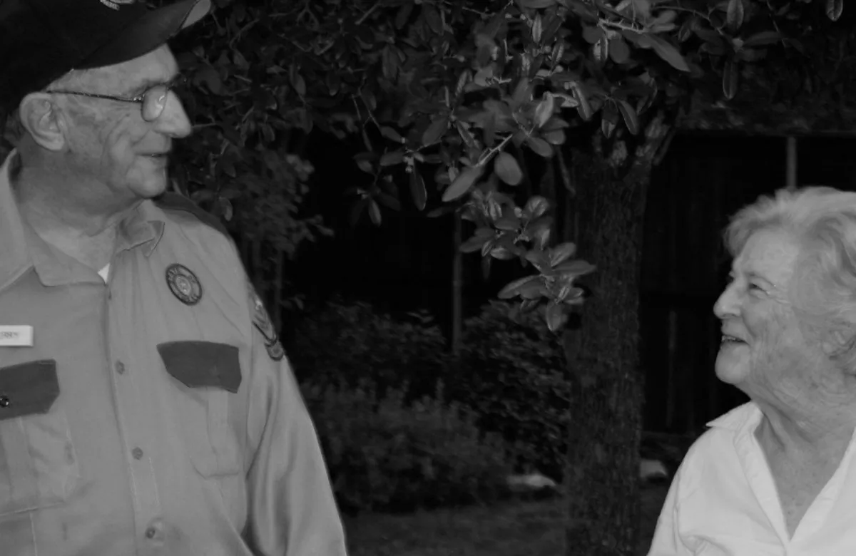 Black and white photo of two elderly people smiling and talking outdoors near trees.