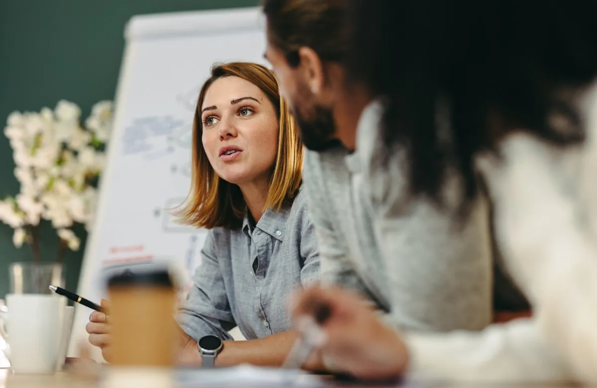 A woman in a gray shirt speaking with two colleagues in a meeting, near a whiteboard.