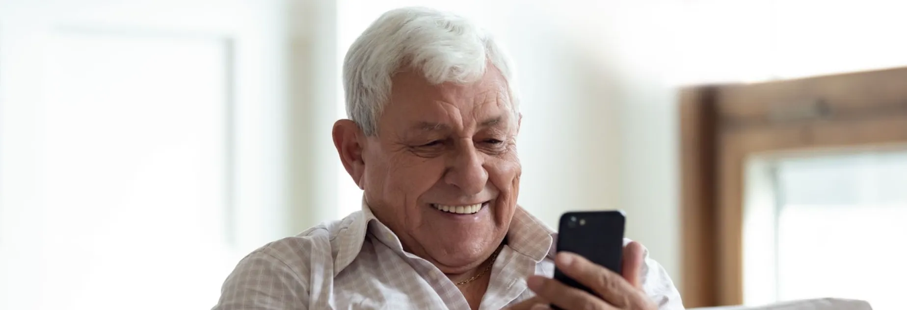 Smiling elderly man looking at smartphone while sitting indoors near a window.