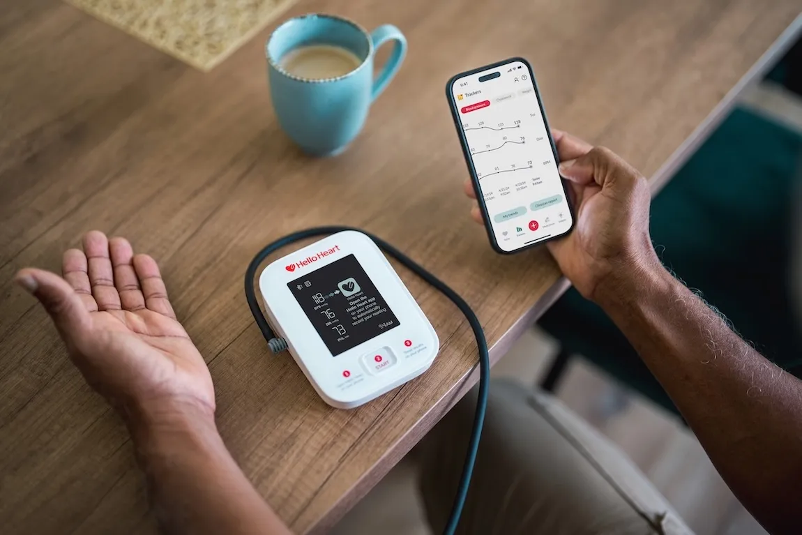 Person using Hello Heart blood pressure monitor with app showing heart health data on phone.