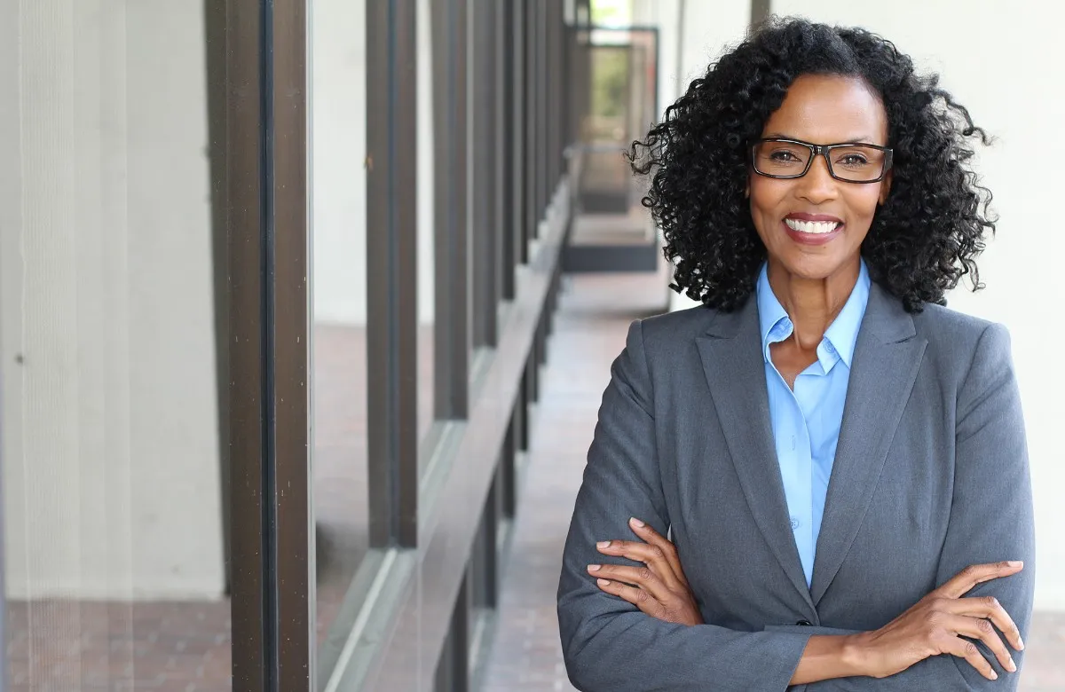 Confident woman in gray blazer and glasses smiling with arms crossed near building windows.