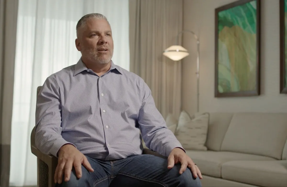 Middle-aged man with gray hair and beard sitting in a chair, speaking in a modern living room.