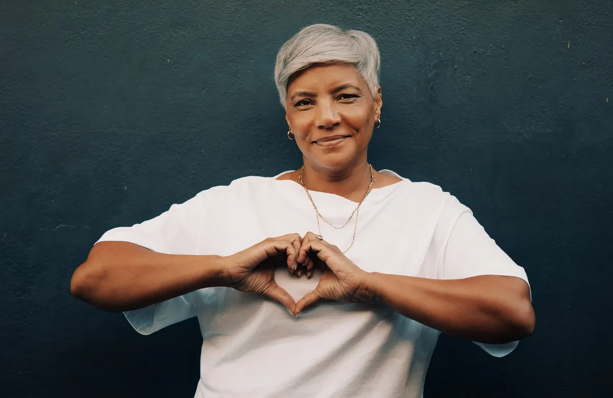 Woman with short gray hair wearing white shirt forming a heart with her hands.