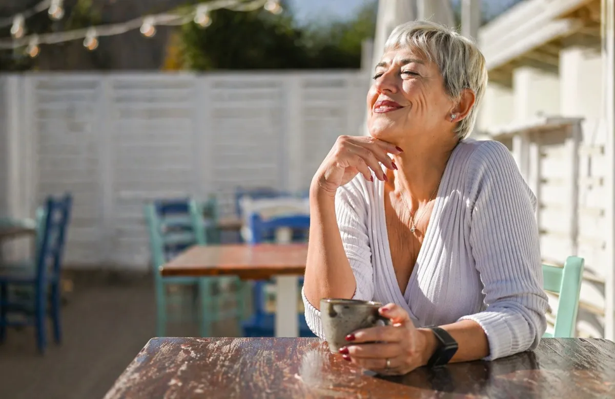 Smiling older woman with short gray hair enjoying a drink at an outdoor cafe table in sunlight.