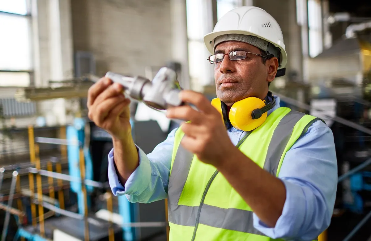 Manufacturing worker wearing a hard hat and safety vest inspects a metal part in a factory.