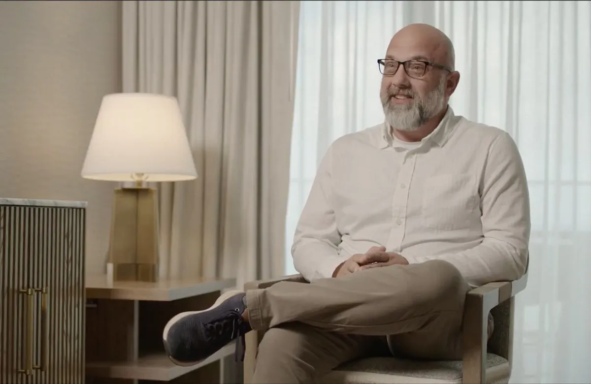 Bald man with glasses and beard sits cross-legged in a chair, smiling, against curtains and lamp.