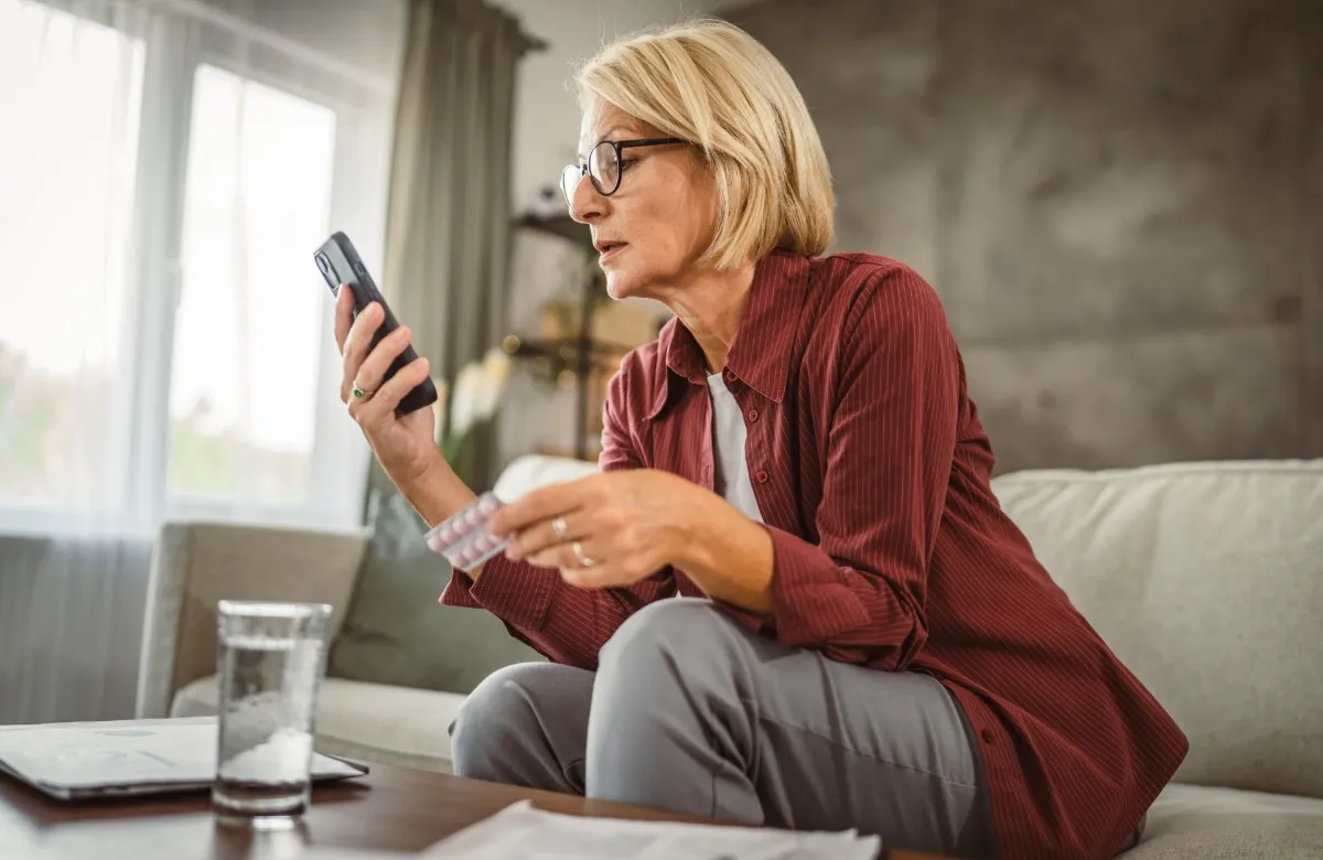 Middle-aged woman holding medication and looking at her phone while sitting on a couch.