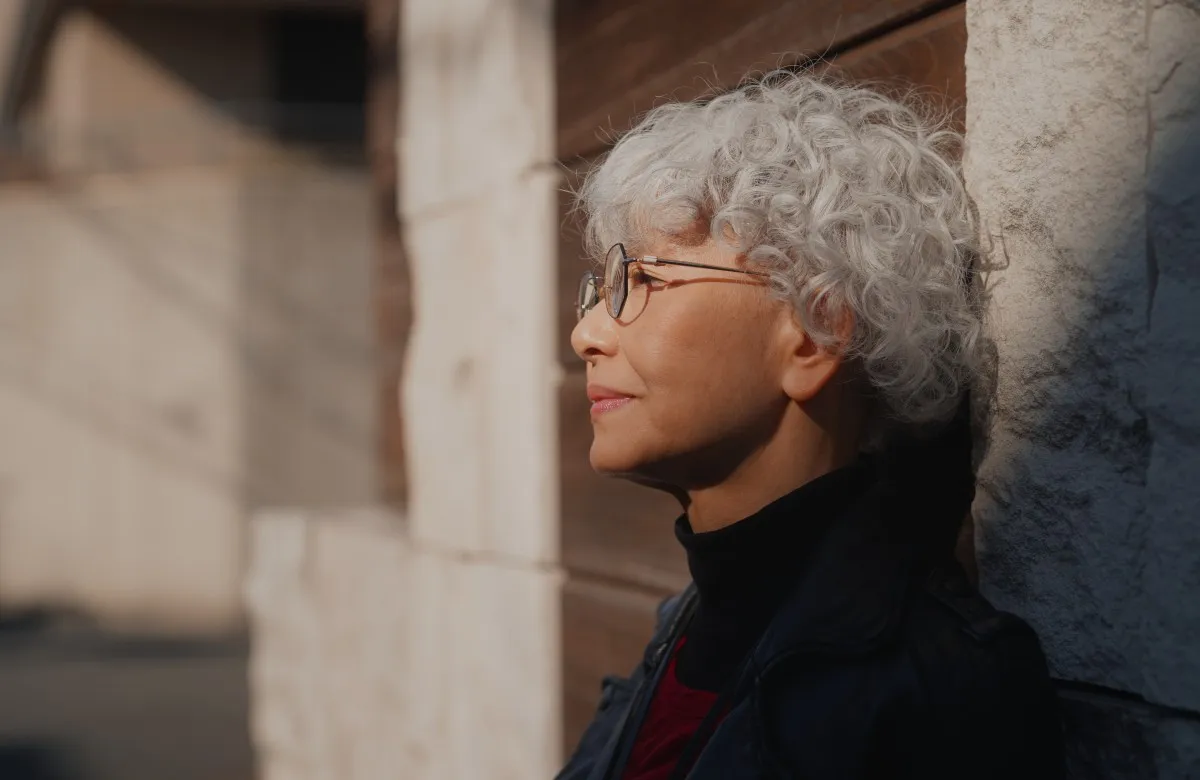 Side profile of a thoughtful older woman with curly white hair and glasses leaning against a stone wall.