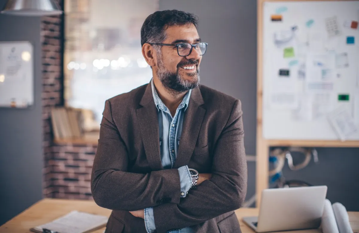 Smiling man with glasses and beard crosses arms in an office with a laptop and whiteboard behind him.
