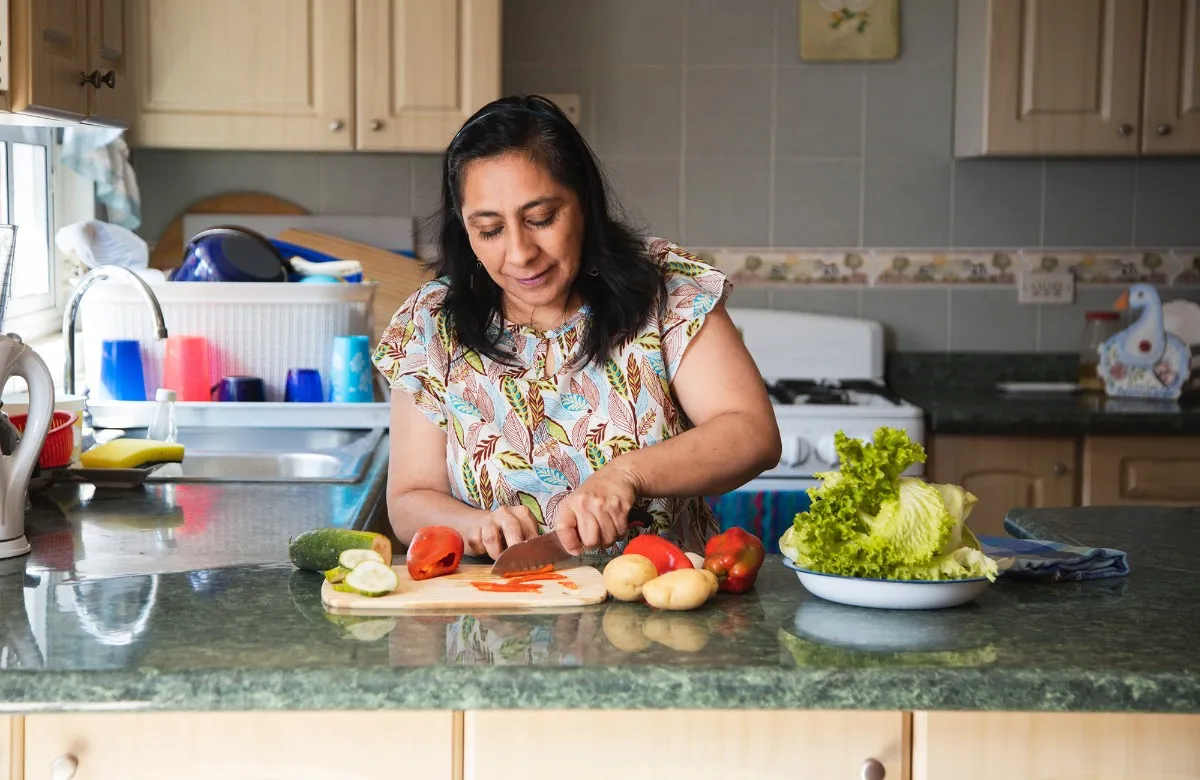 Woman in a kitchen chopping red bell pepper on a cutting board with vegetables around her.