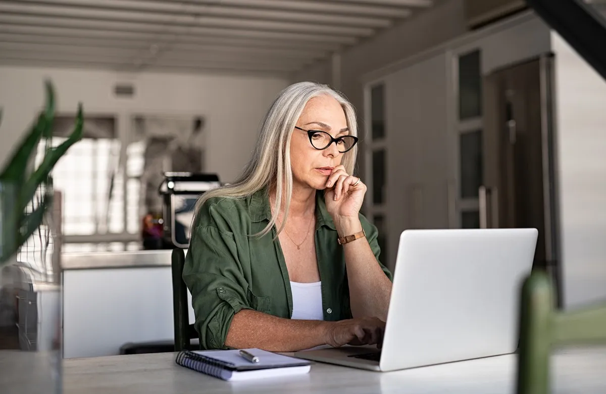 Woman with long gray hair and glasses working on a laptop at a table with a notebook and pen.