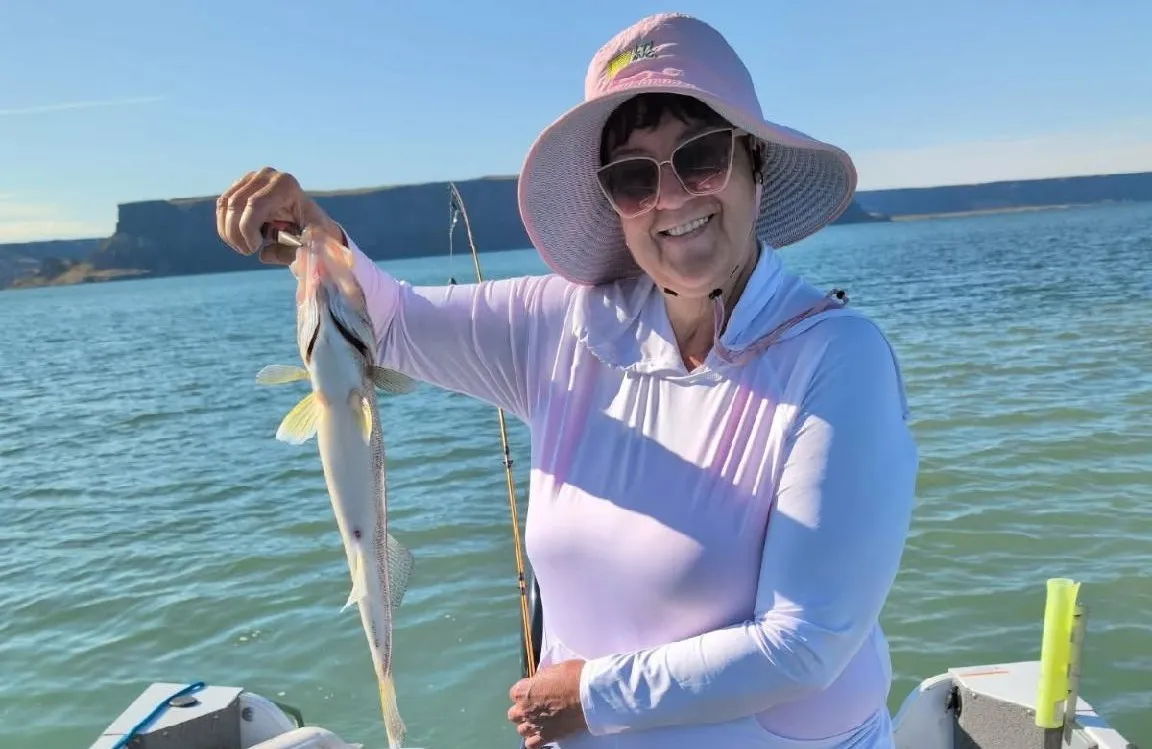 Smiling woman in sunhat and sunglasses holding a fish on a boat with water and cliffs in the background.