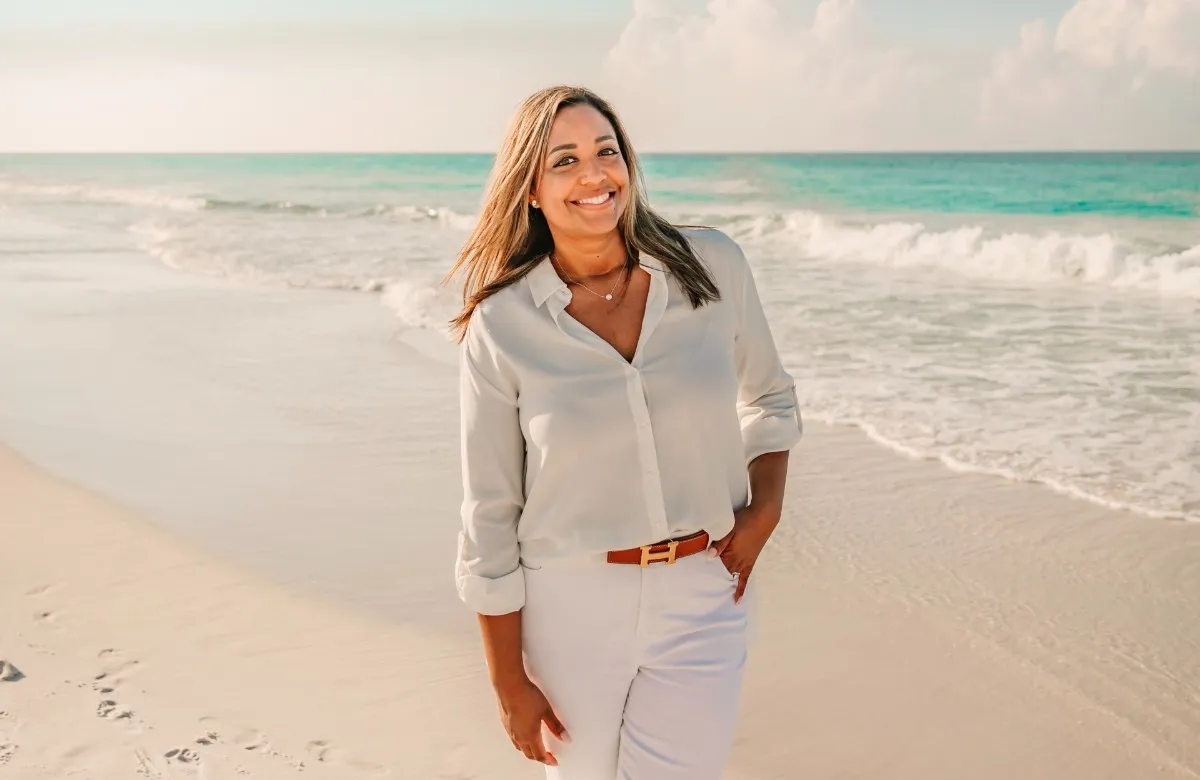 Smiling woman in white blouse and pants standing on a sunny beach with waves in the background.