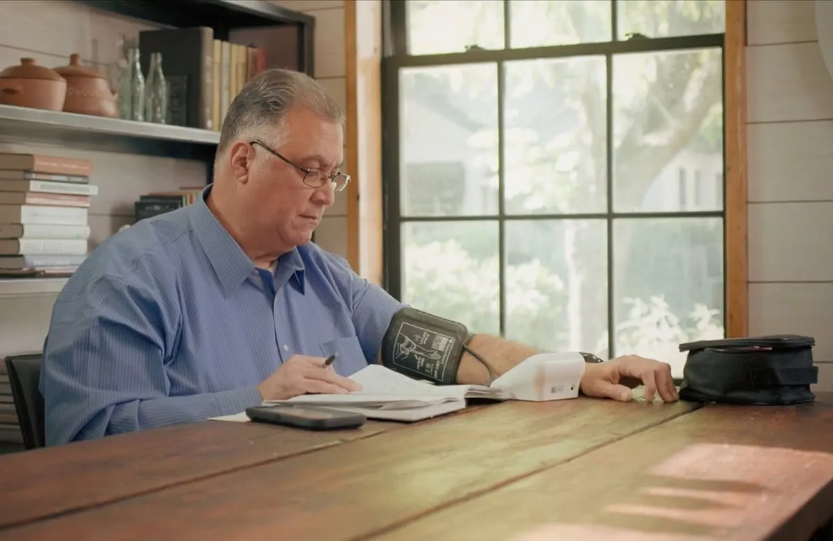 Man in glasses and blue shirt taking blood pressure while writing notes at wooden table.