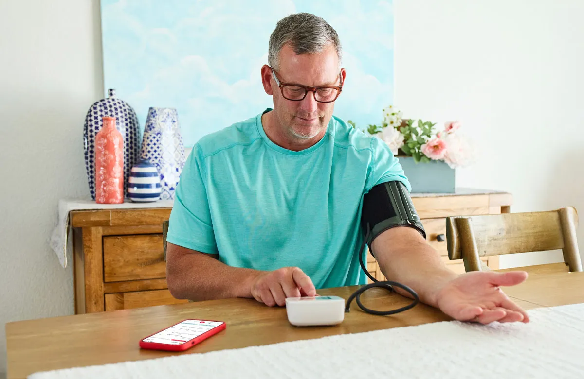 Man in turquoise shirt checking blood pressure at home with monitor and smartphone on table.