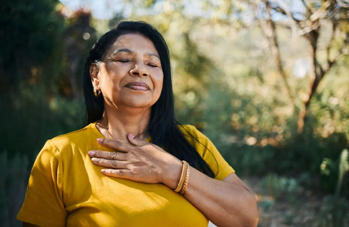 Woman with eyes closed and hand on chest wearing yellow shirt outdoors with green blurred background.