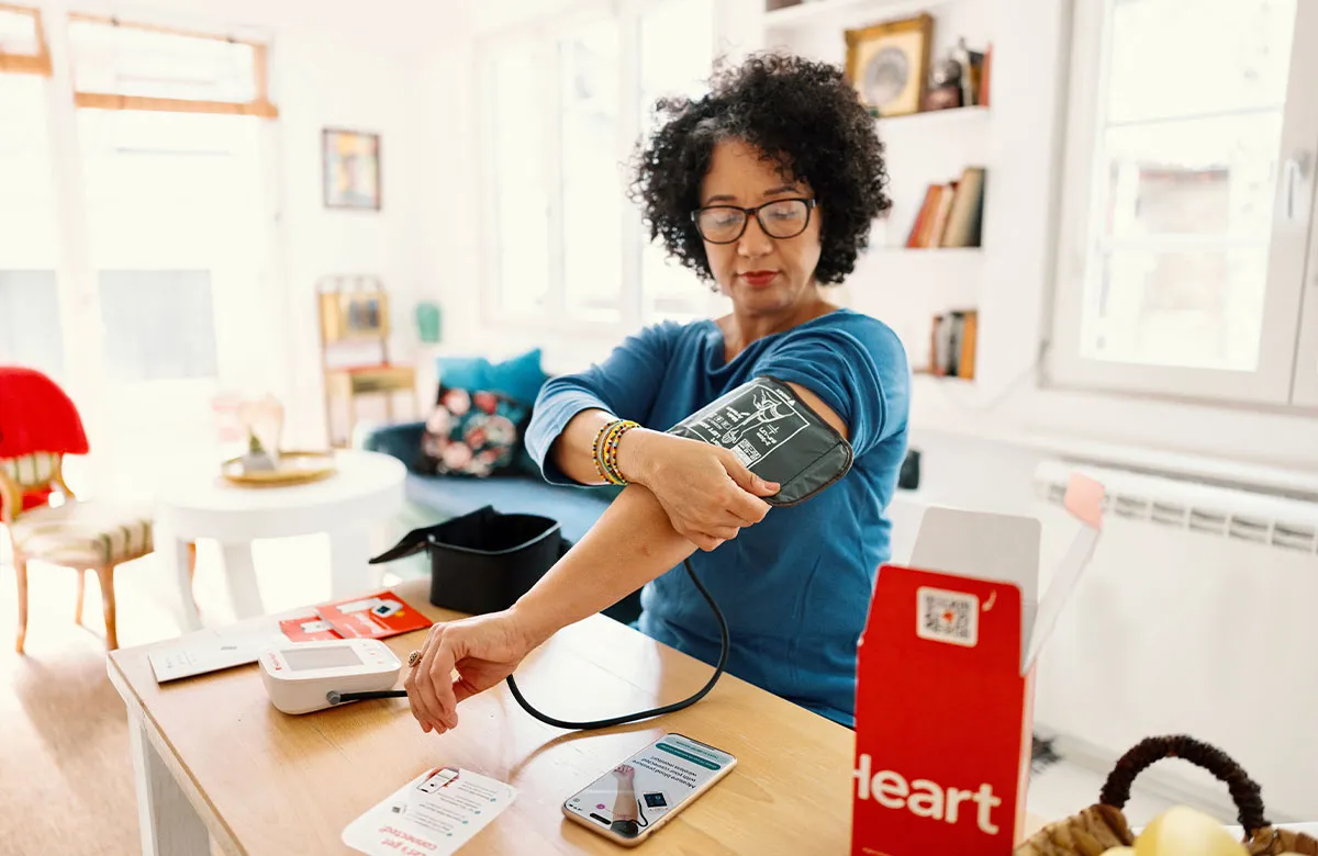 Woman using digital blood pressure monitor at home, with smartphone showing related app on a table.