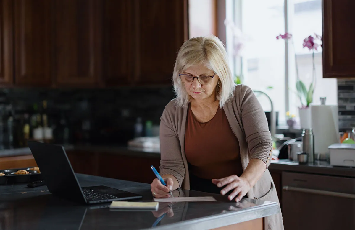 Woman with blonde hair and glasses writing on paper at a kitchen counter with an open laptop nearby.