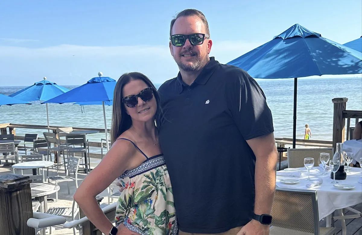 Man and woman wearing sunglasses pose smiling at a seaside restaurant with blue umbrellas and ocean view.
