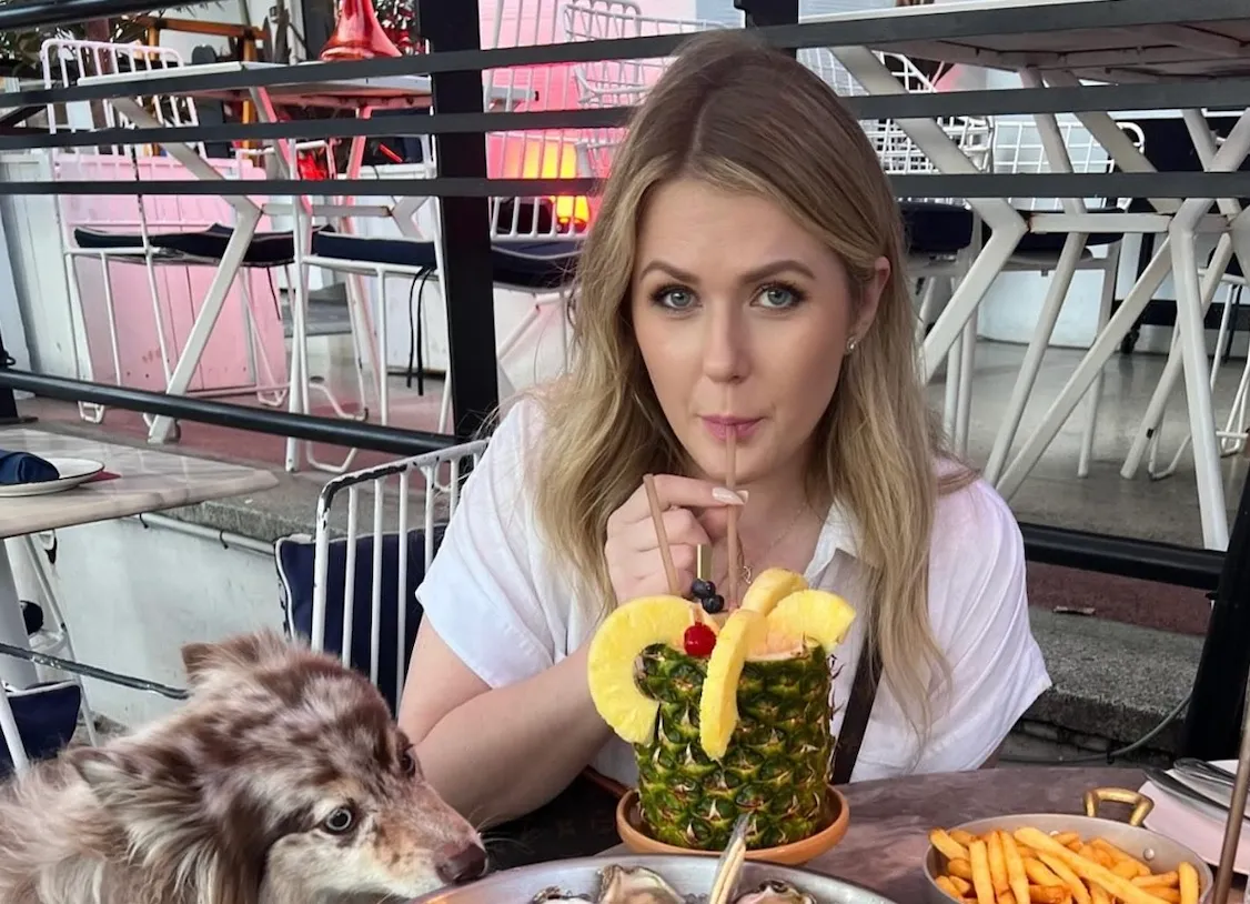Woman in white shirt sipping a pineapple drink at a table with a dog and food.