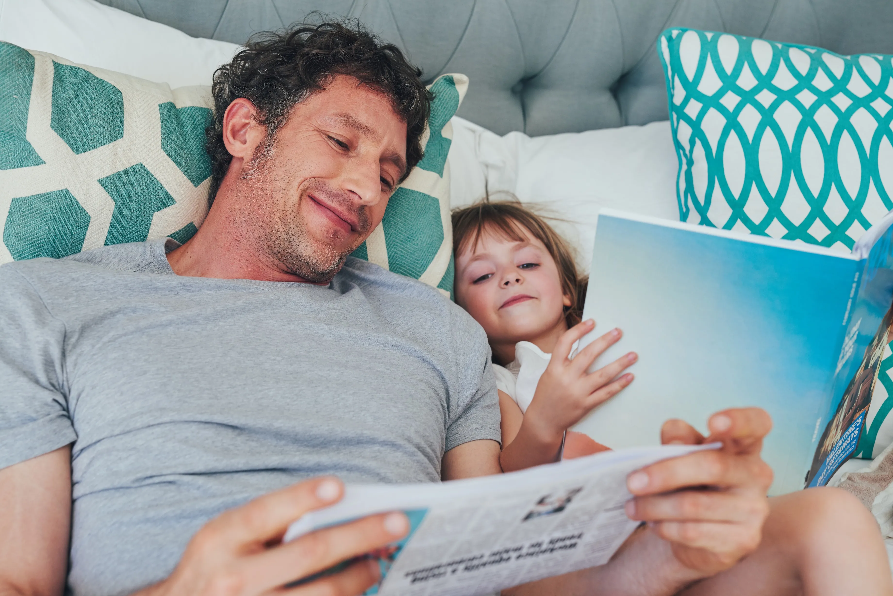 Father and young daughter reading books together lying on a bed with patterned pillows.
