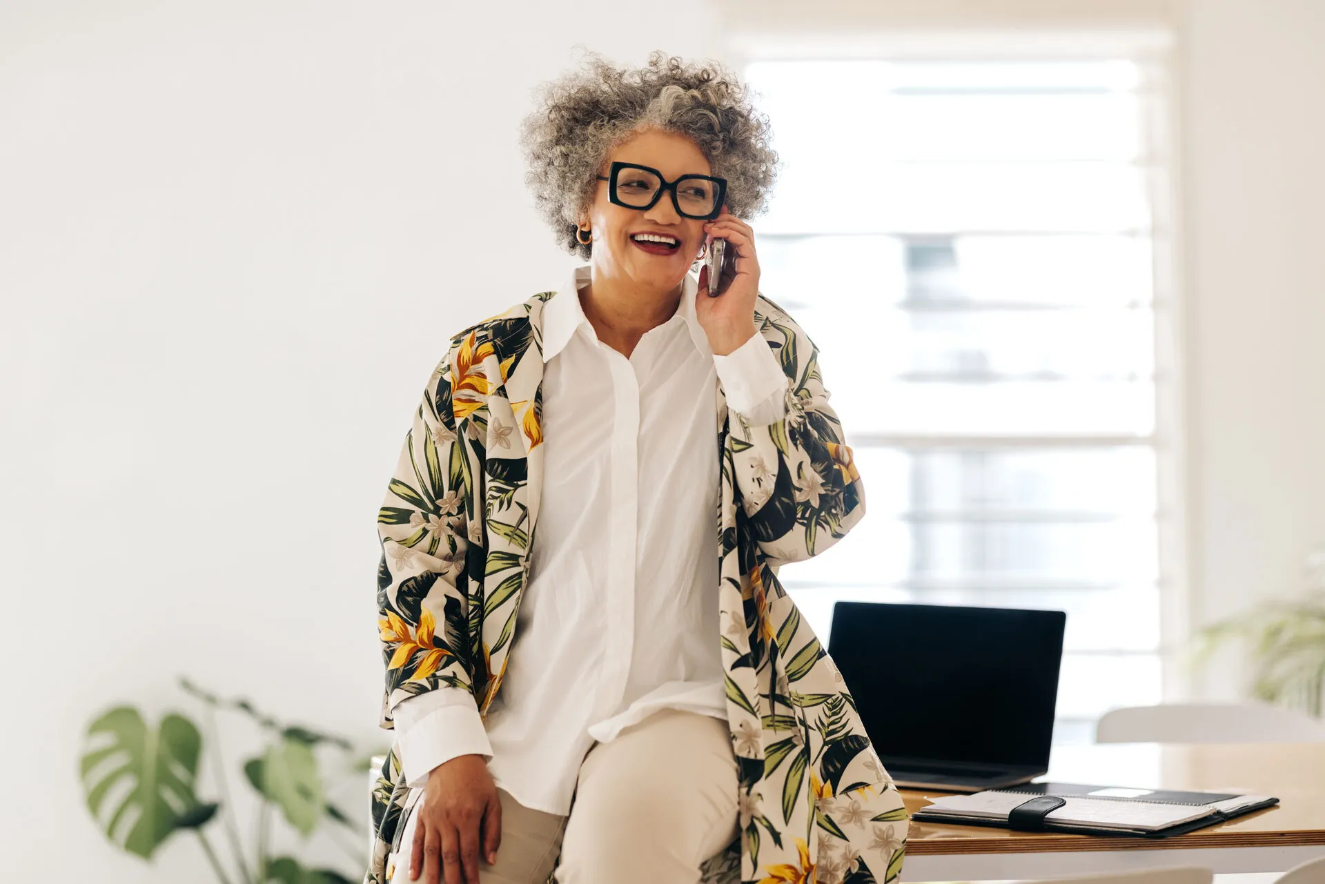 Smiling woman with curly gray hair talking on phone, sitting on desk with laptop and planner.