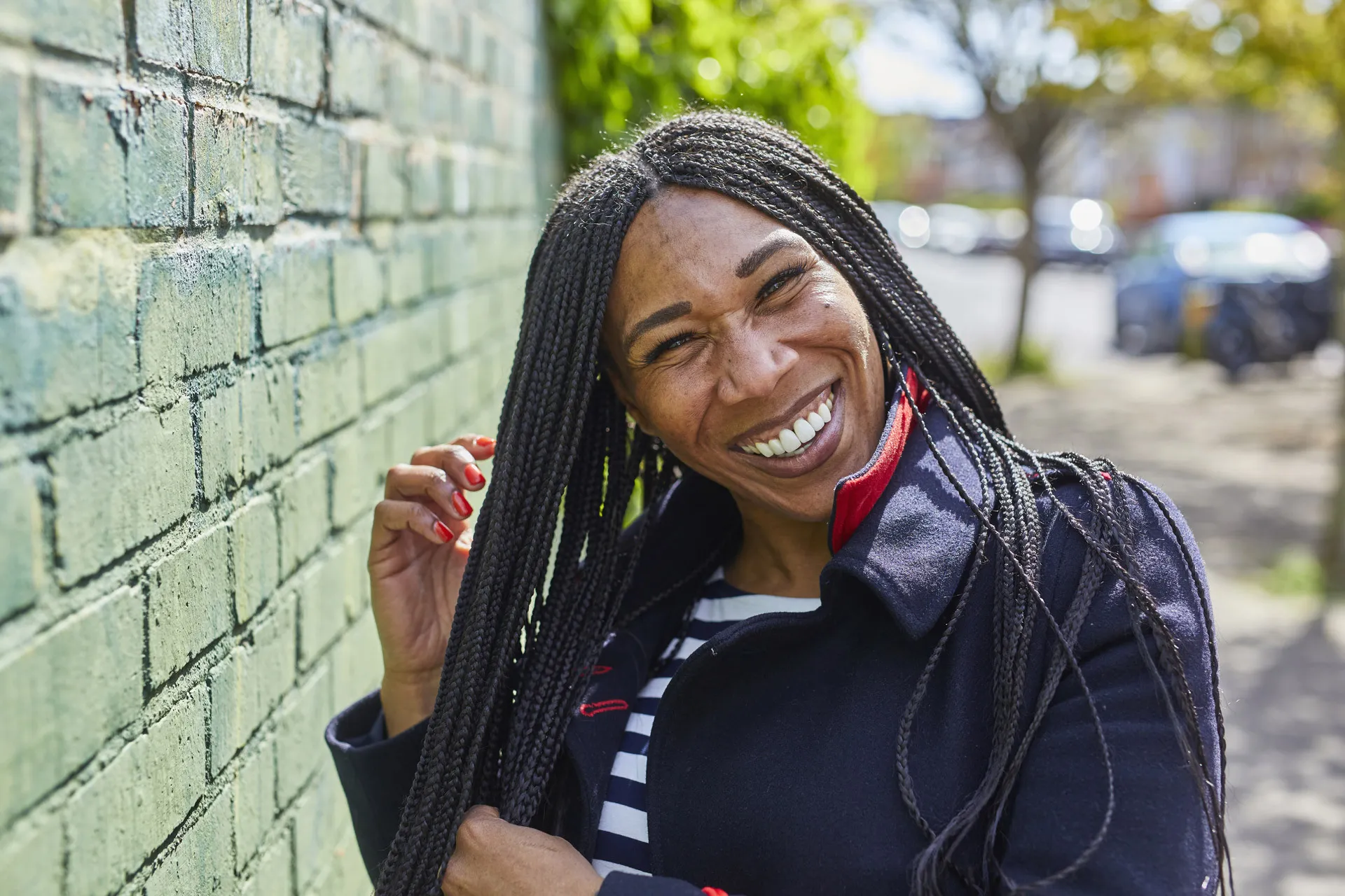 Smiling woman with long braided hair wearing a navy coat and striped shirt near a green brick wall.