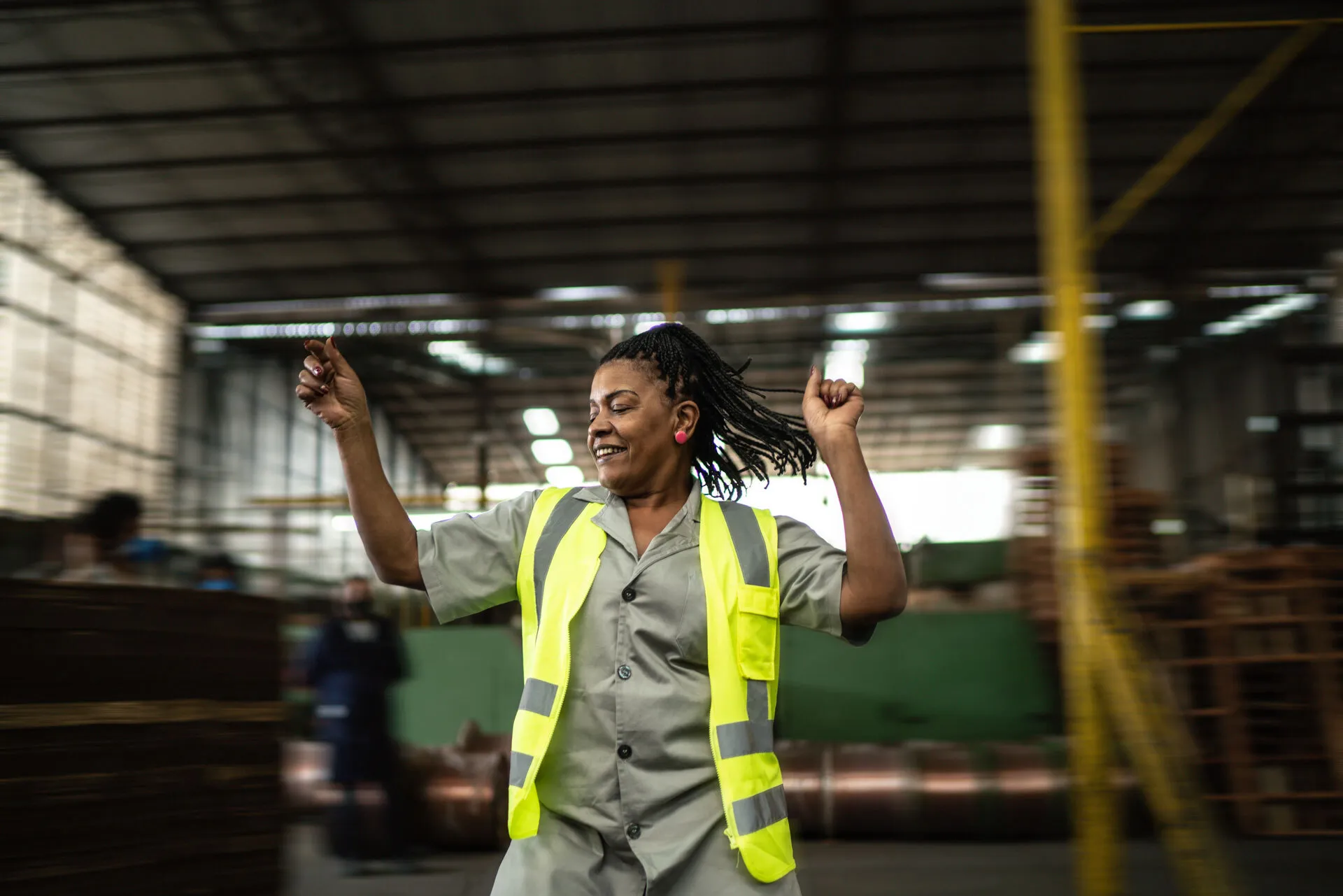 Woman in safety vest dancing joyfully in an industrial warehouse setting.