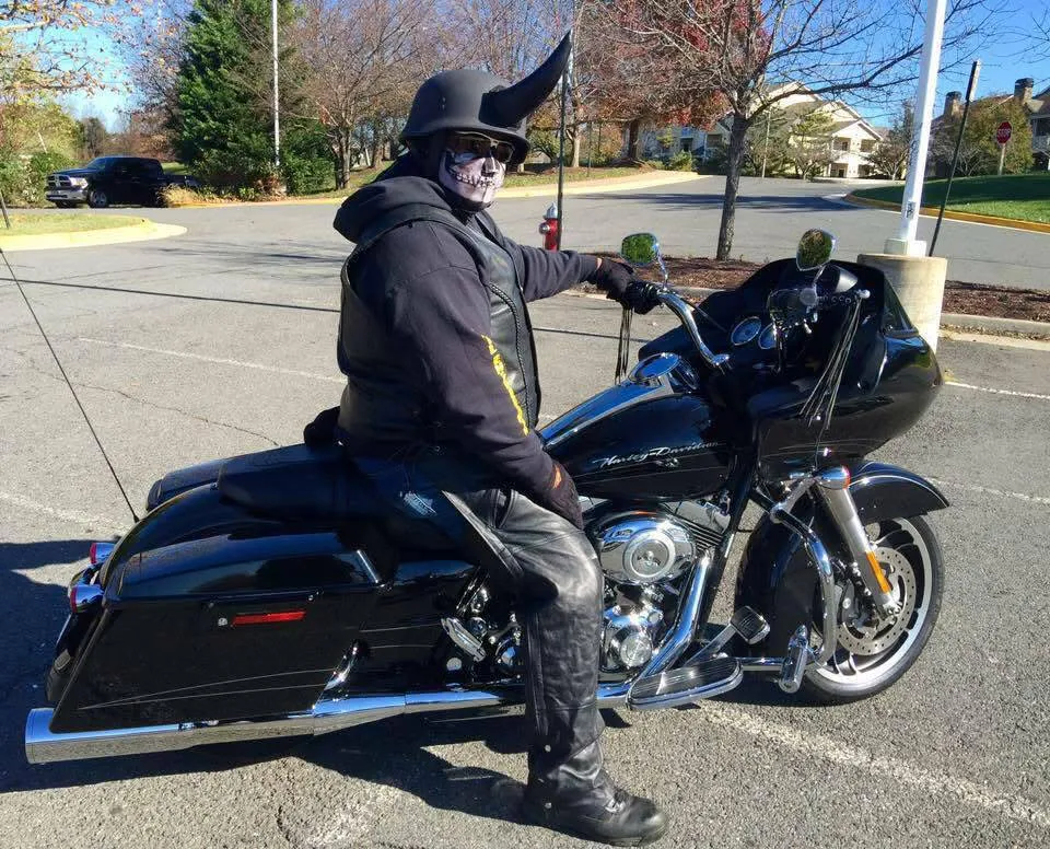 Person in skull mask and horned helmet sitting on black Harley-Davidson motorcycle in a parking lot.