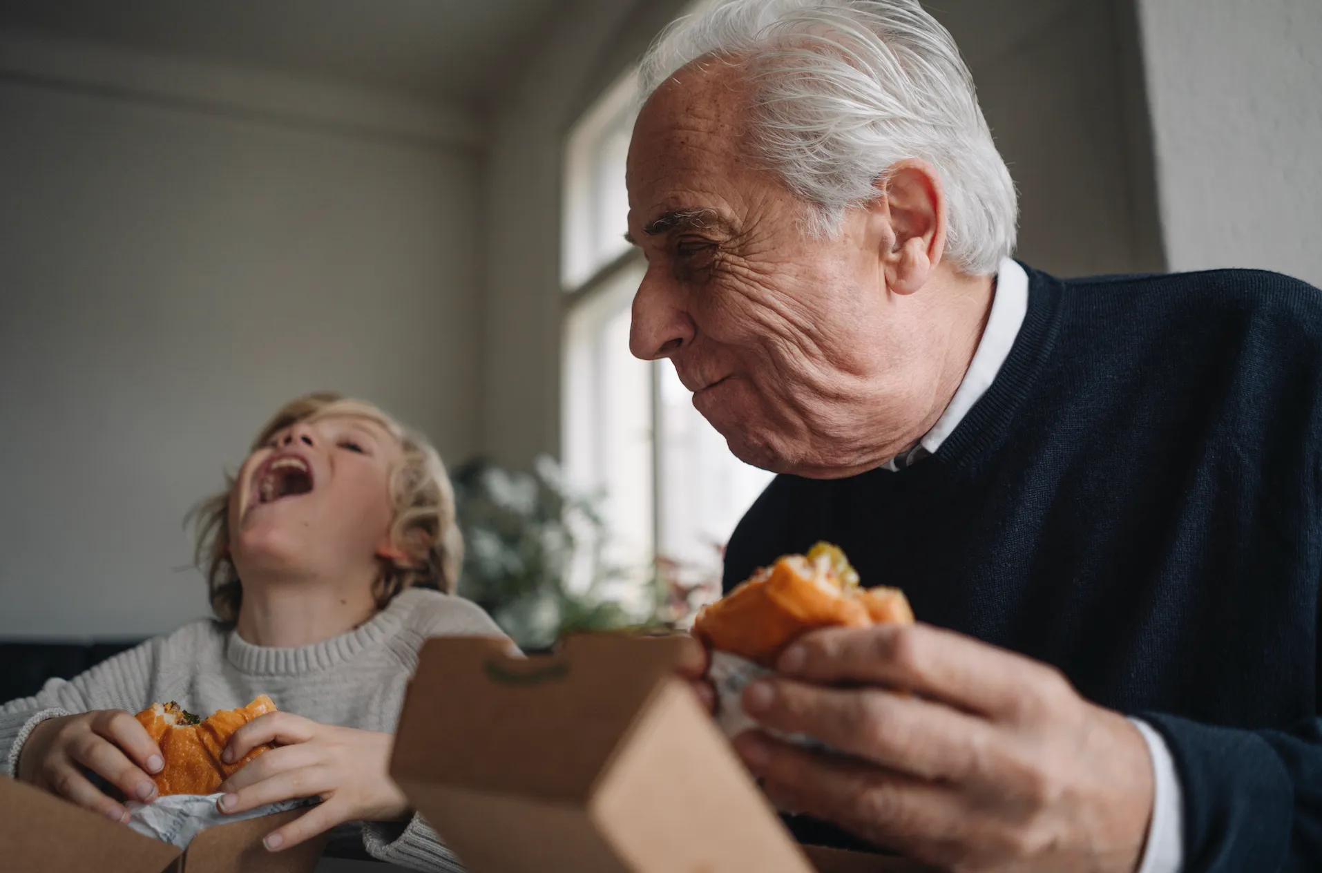 An elderly man and child laugh together while holding sandwiches inside a home.