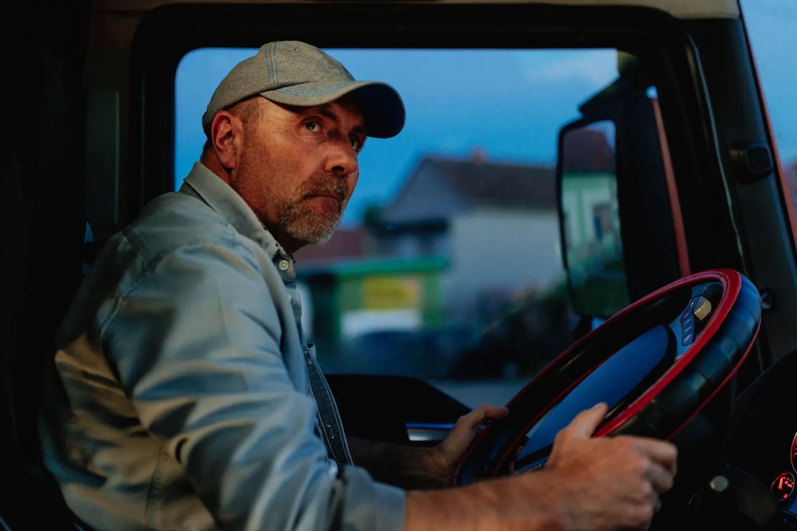 Man with gray cap driving a vehicle at dusk, holding a steering wheel with red and black cover.