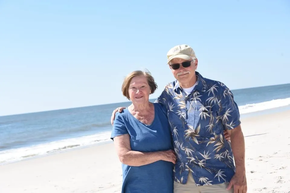 Older couple standing arm in arm on a sunny beach with ocean waves in the background.