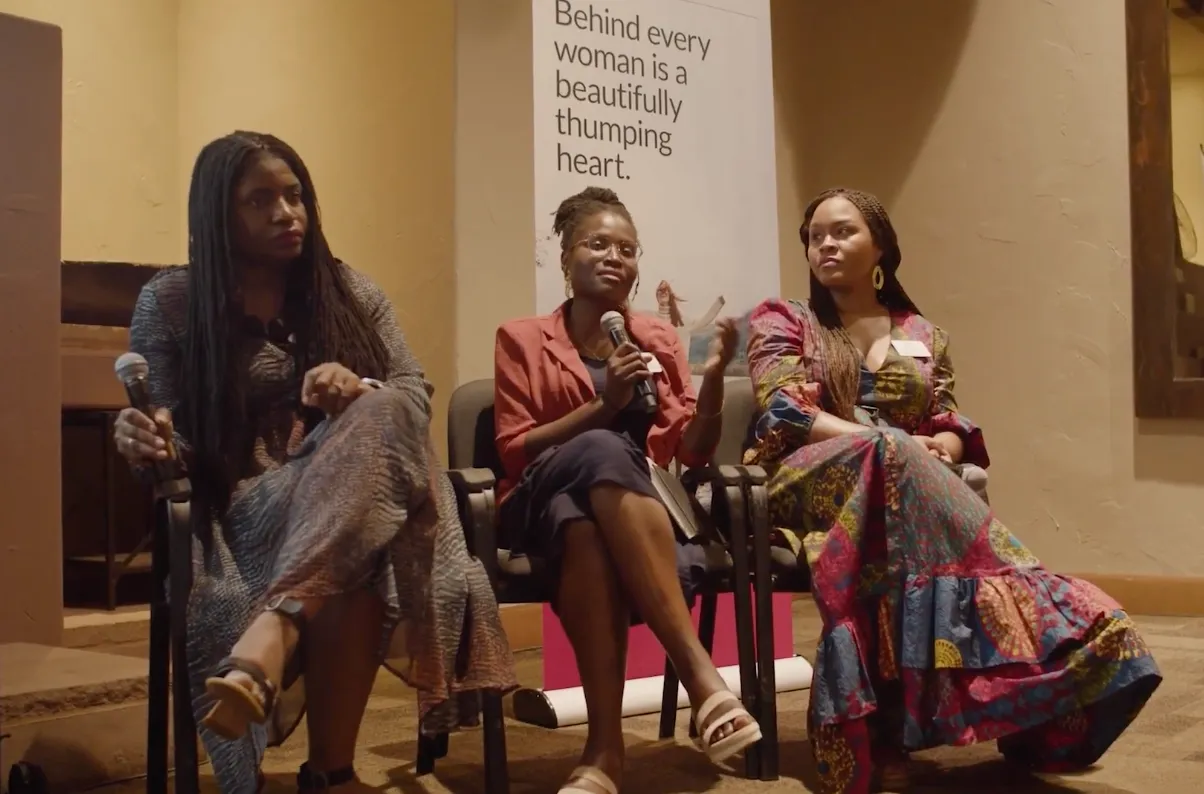 Three women seated on chairs, one speaking with a microphone, with a poster behind them.