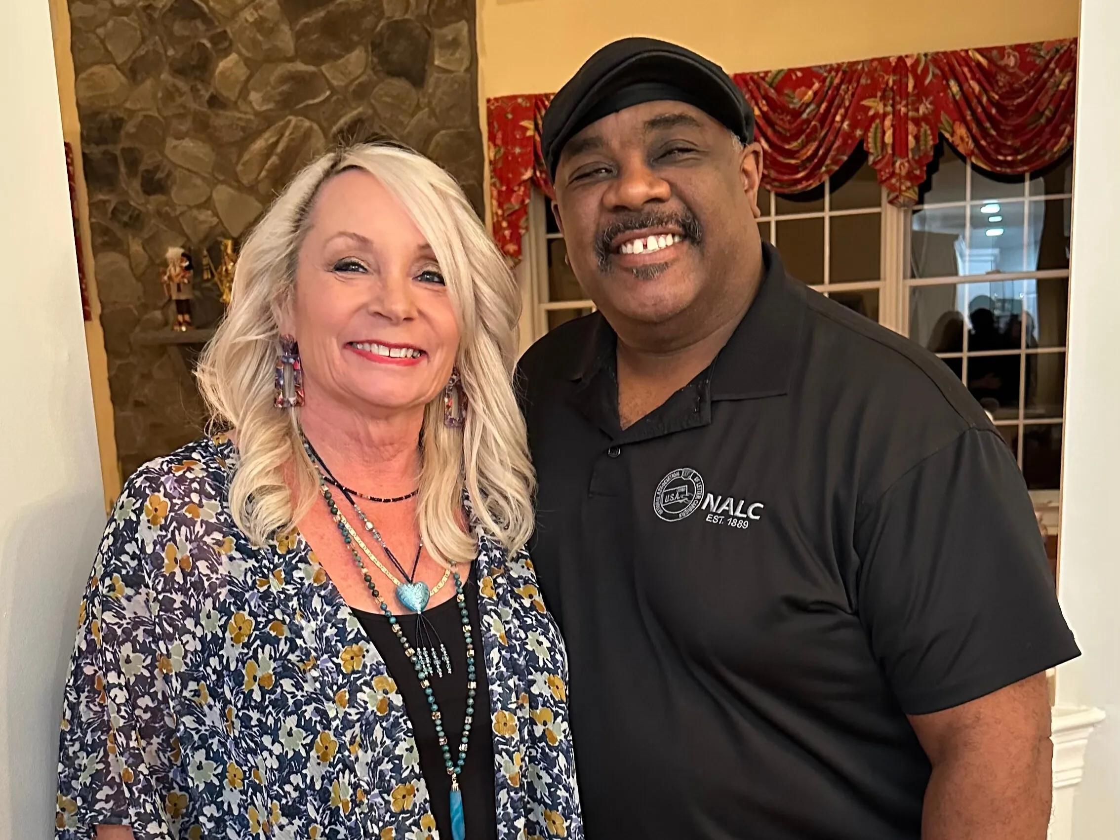 Smiling woman with blonde hair and floral top posing with man in black shirt and cap indoors.