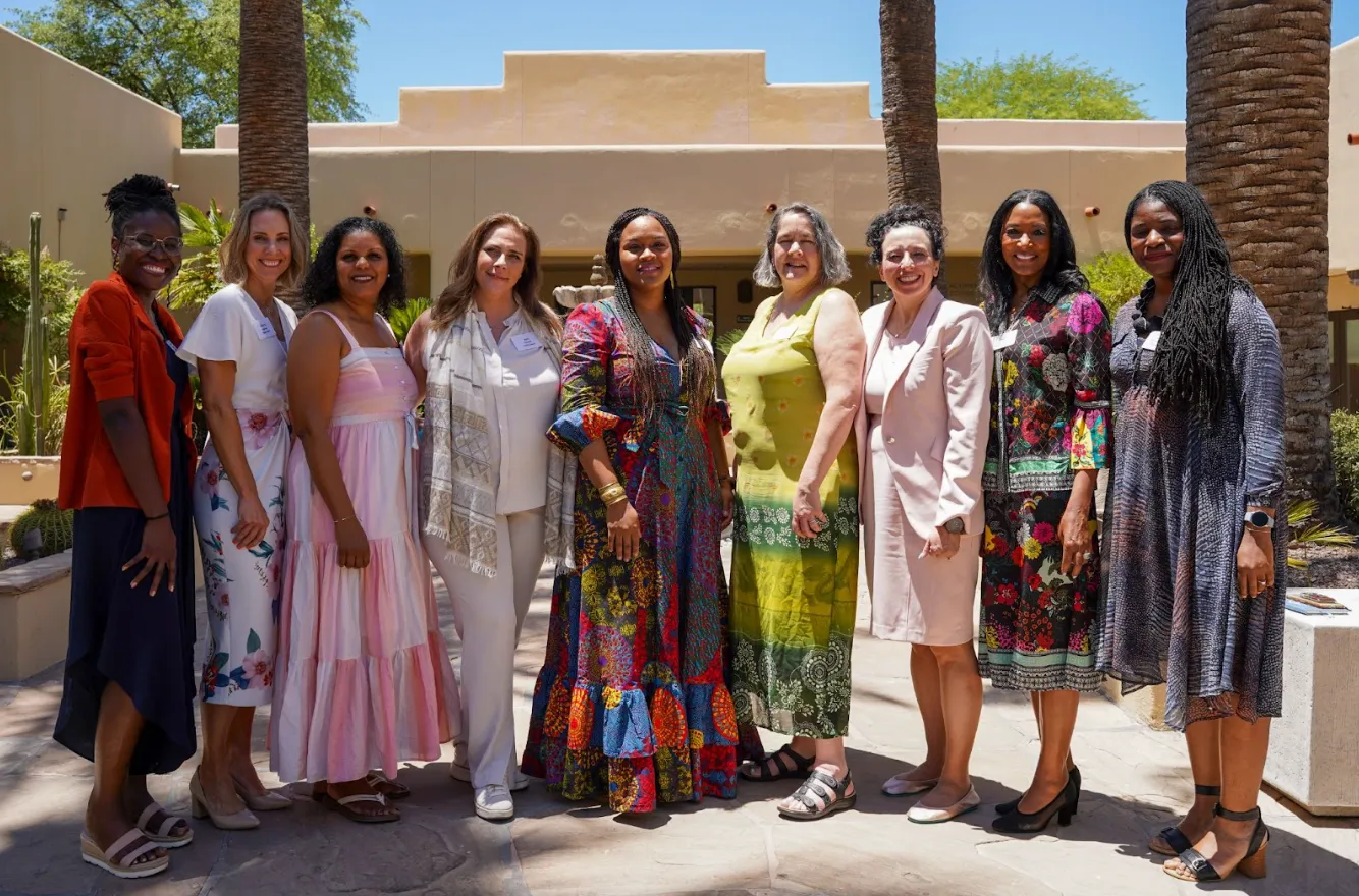 Group of nine women standing outdoors in a sunny courtyard with a fountain and palm trees.