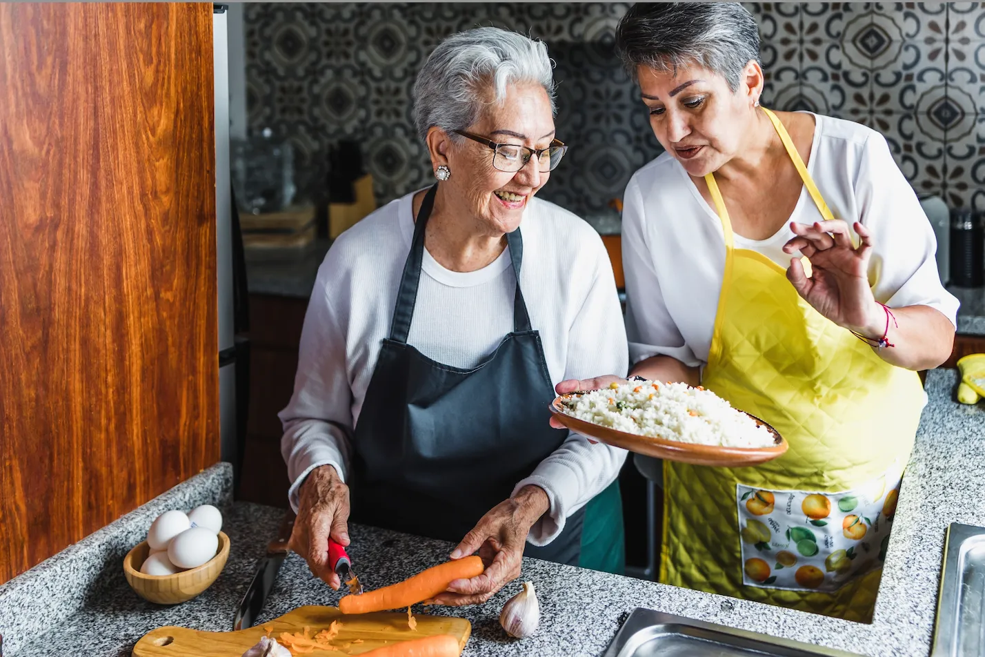 Two women preparing food in kitchen; one peeling a carrot, the other holding a plate of rice.
