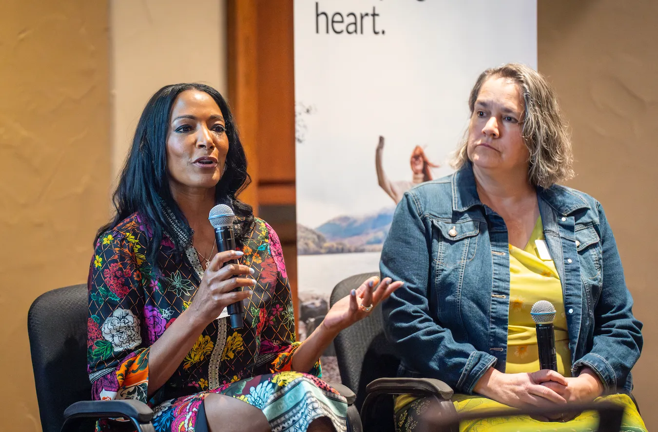 Two women seated speaking during a panel; one holds a microphone and gestures while the other listens.