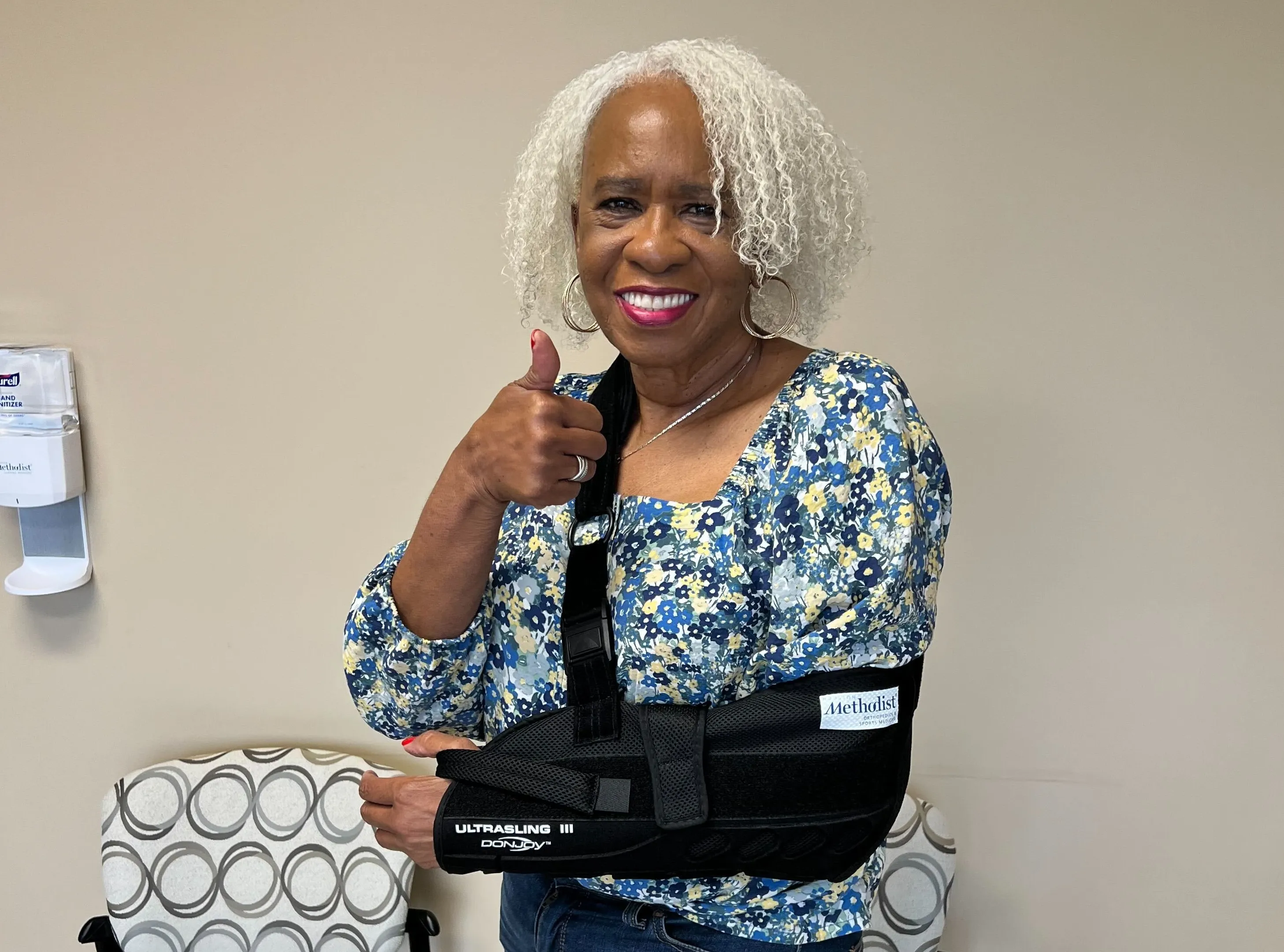 Smiling older woman wearing arm sling giving thumbs up in a medical setting.
