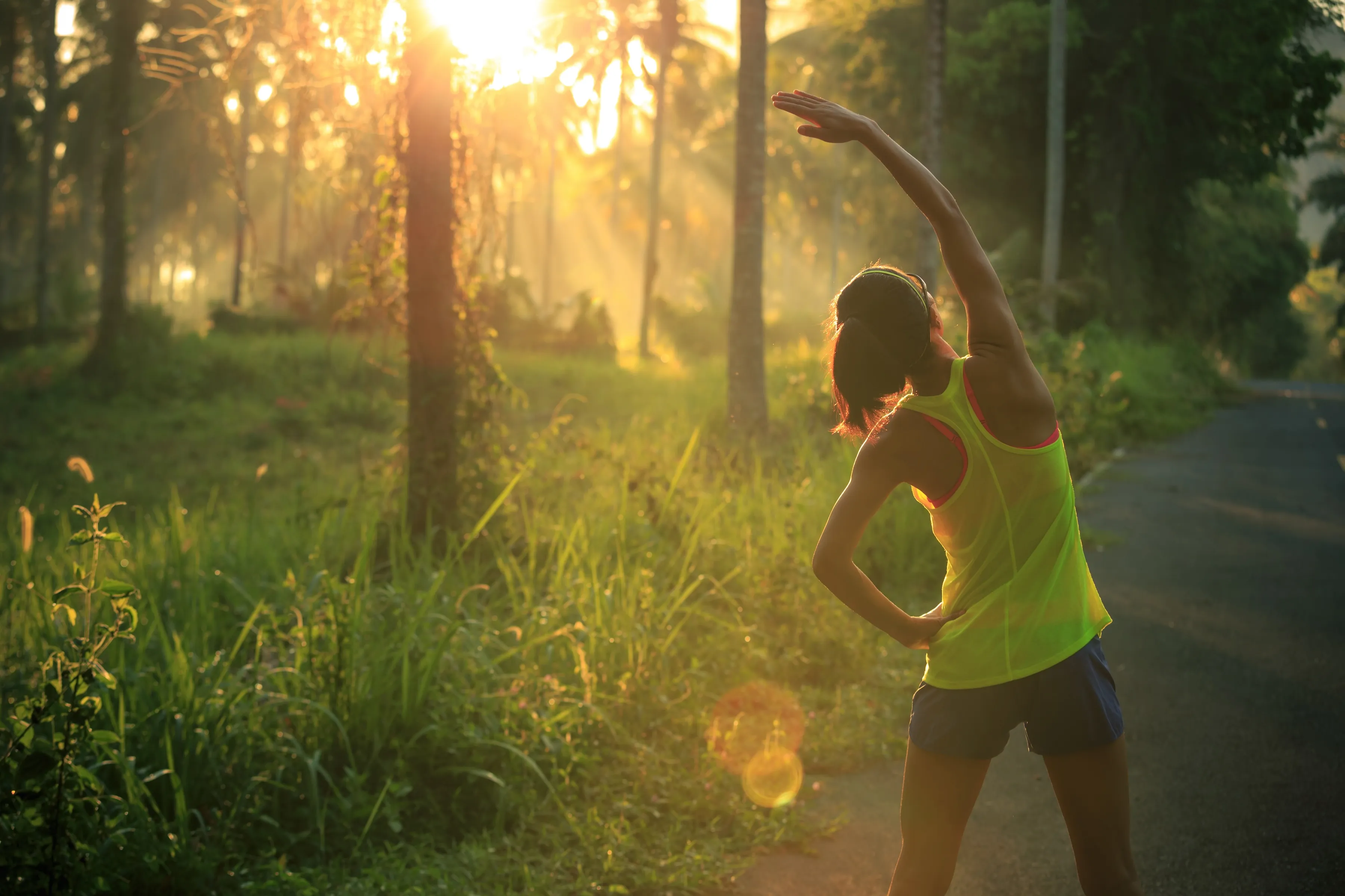 Woman stretching beside a road in a sunlit forest clearing during sunrise or sunset.