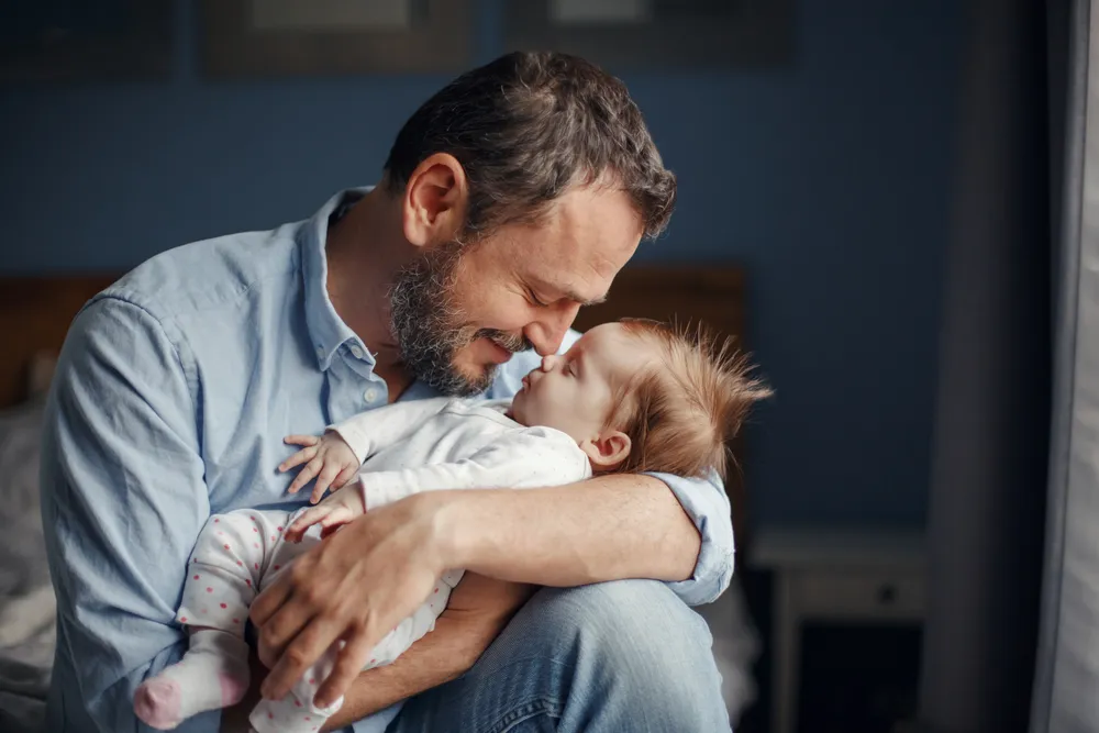 Father wearing blue shirt holding and gently touching noses with a baby in a white outfit.