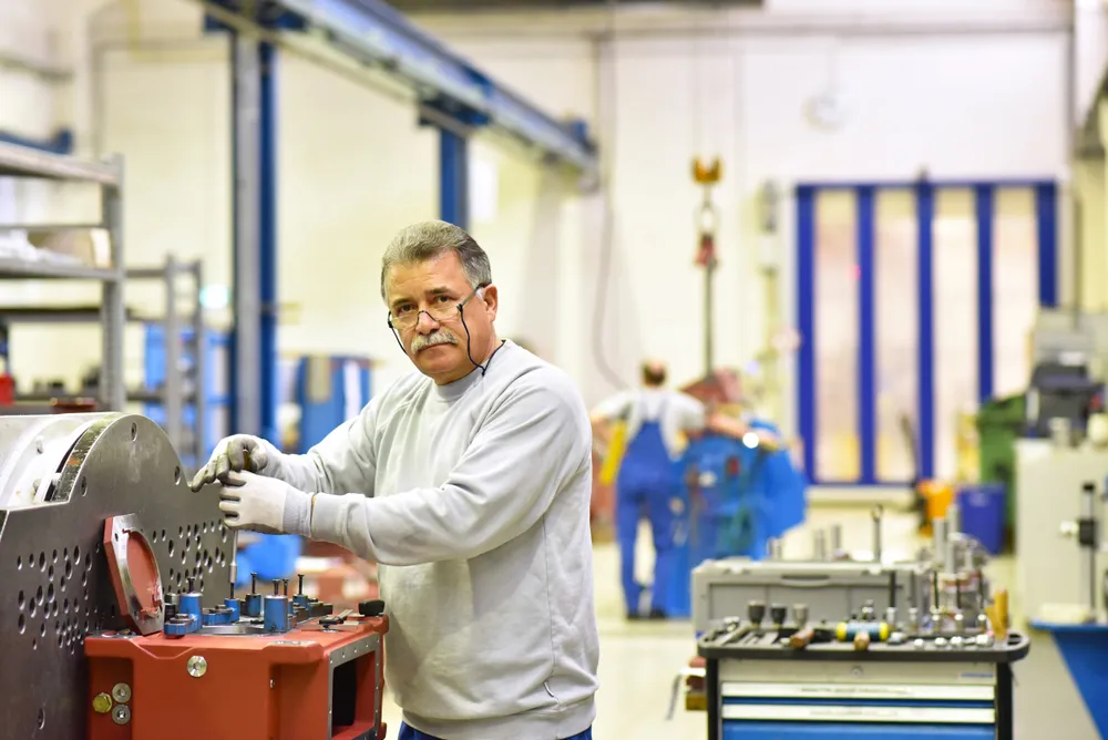 Man in light gray sweatshirt and glasses works with machinery in a factory setting.
