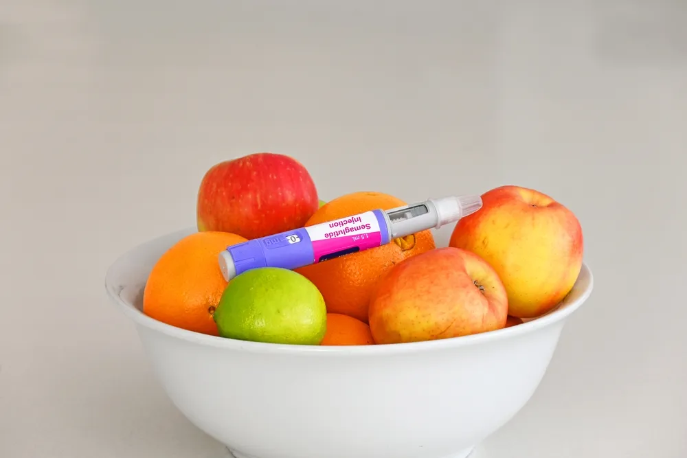 White bowl with apples, oranges, a lime, and a blood glucose test pen on top of the fruit.
