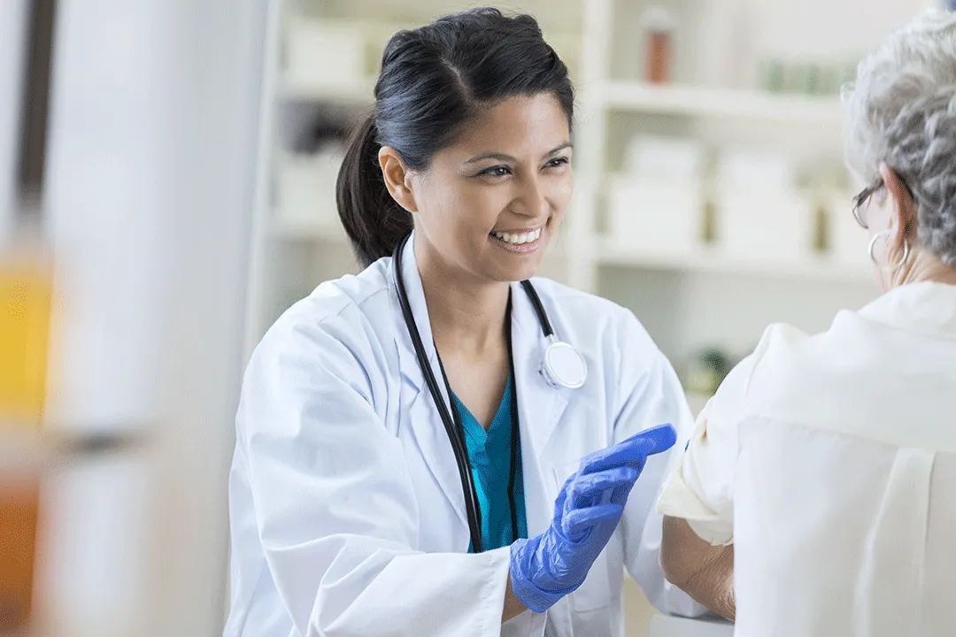 Female doctor with stethoscope and gloves smiling while preparing to give an elderly patient an injection.