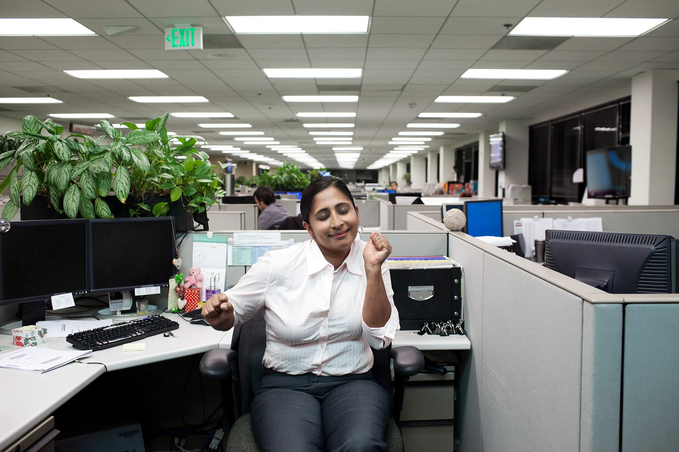 Woman in office cubicle stretching and smiling with eyes closed amid row of workstations.