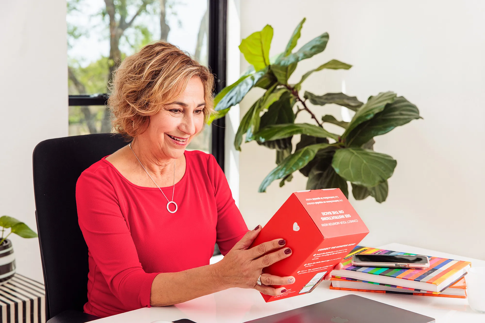 Smiling woman in red shirt holding a red box, sitting at a desk with plant and notebooks behind.