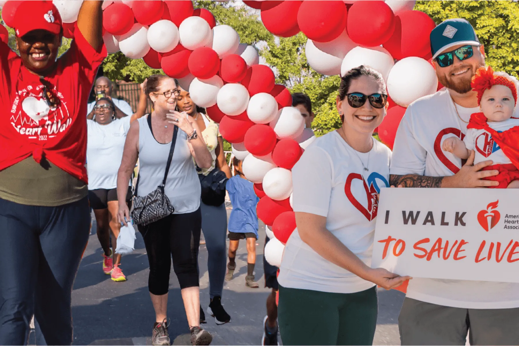 Smiling diverse group walking outdoors under red and white balloon arch for heart health awareness event.