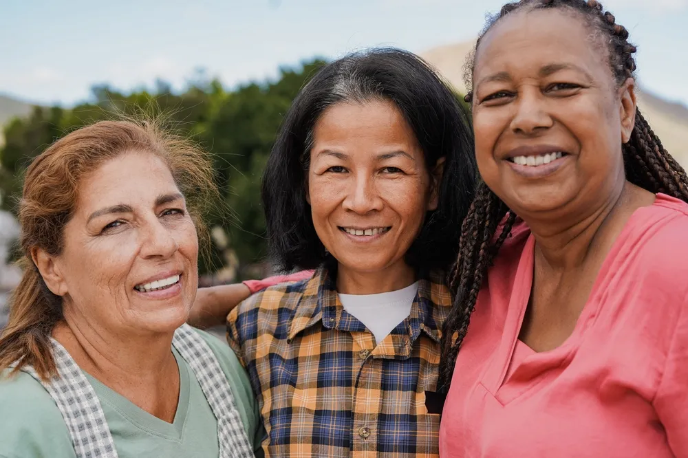Three smiling middle-aged women posing outdoors with trees and sky in the background.