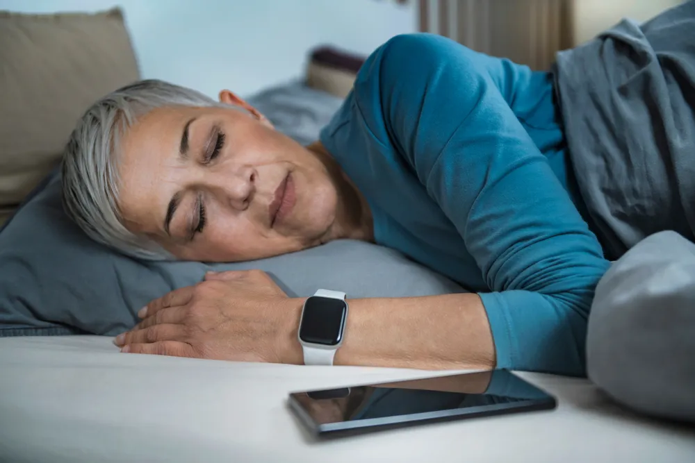 Older woman with short gray hair sleeping on a bed wearing a blue shirt and smartwatch, phone nearby