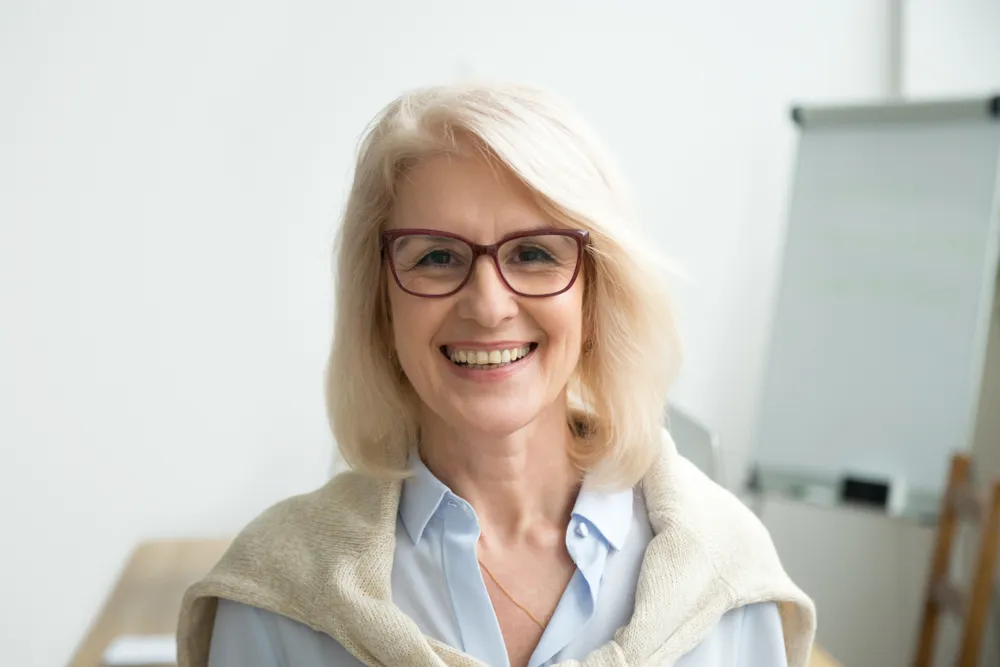 Smiling middle-aged woman with blonde hair and glasses wearing a light sweater over her shoulders.